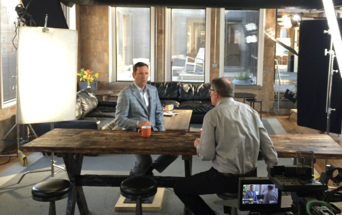 Location shot with big windows. Show two men sitting at a long wooden table in interview setting. Big lights surround the shoot setting