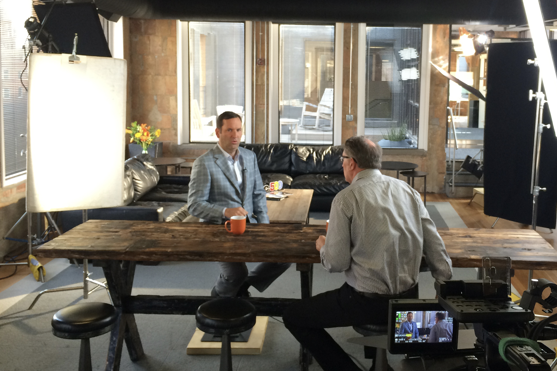 Location shot with big windows. Show two men sitting at a long wooden table in interview setting. Big lights surround the shoot setting